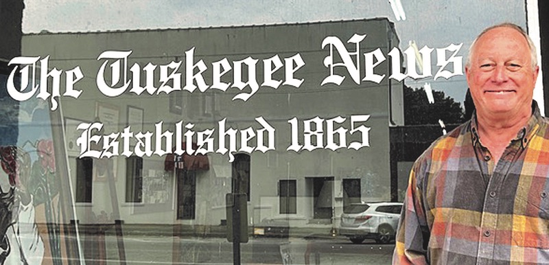 Joe Turnham stands outside a storefront window displaying the name “The Tuskegee News” and the words “Established 1865”. The window reflects buildings, cars, and street signs from across the road.